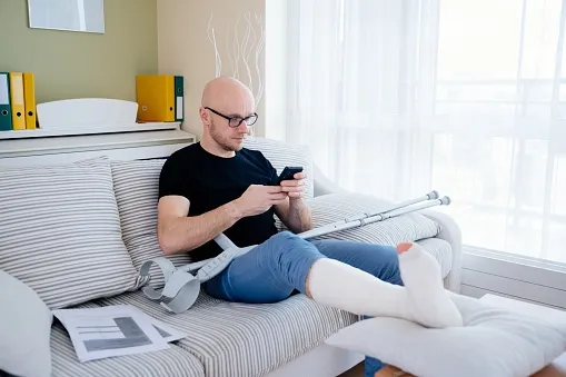 A man in a cast and crutches sitting on a couch texting his attorney.